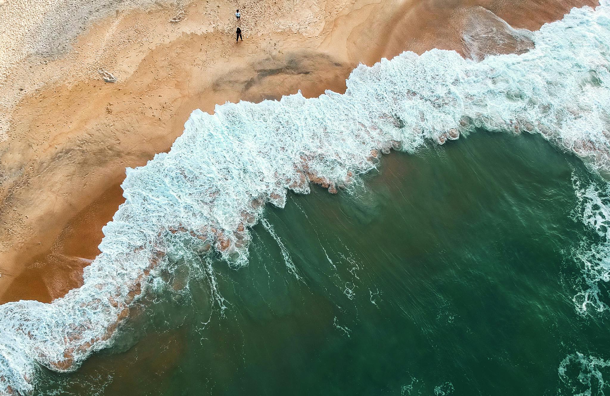 About us Aerial shot of ocean waves crashing onto a sandy beach with two people in the distance.
